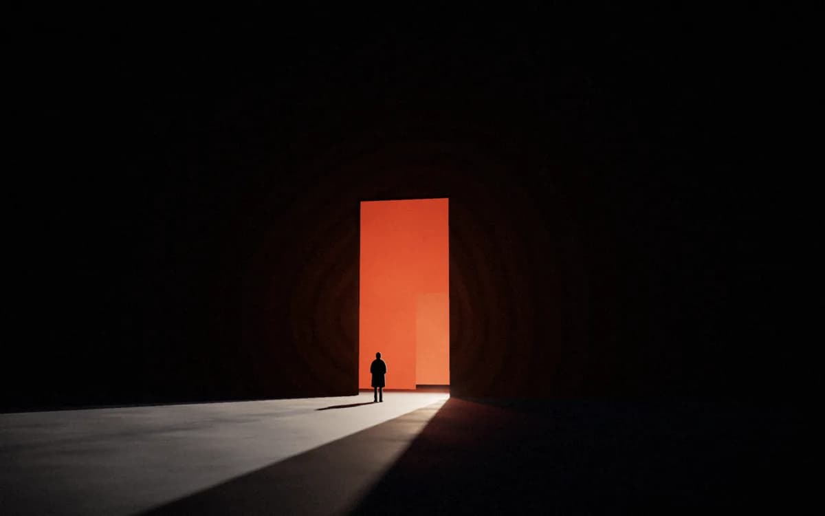 A silhouetted figure standing before a tall doorway of orange light in a black hall, the spill dragging their shadow across a concrete floor